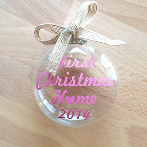 May include: A clear, spherical glass Christmas ornament topped with a natural tan burlap ribbon bow. The front of the ornament features pink lettering that reads First Christmas Home with a heart replacing the letter o, followed by the year 2014.
