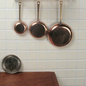 May include: Three copper cooking pans of different sizes hanging on a white tiled wall. The pans have gold handles and are arranged in a row from largest to smallest.