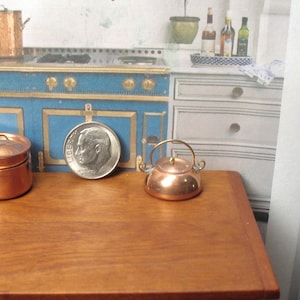 May include: A miniature copper teapot with a gold handle sits on a wooden table next to a copper pot and a US dime.