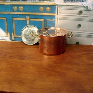 Peut inclure: Une petite casserole en cuivre avec un couvercle et deux poignées. La casserole a une finition martelée et est posée sur une surface en bois.
