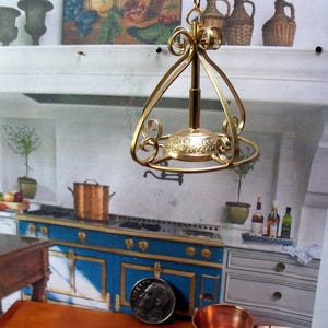 May include: A gold-colored, ornate miniature chandelier with a decorative base and scrolled accents. The chandelier is suspended from a chain and is designed for a dollhouse or miniature setting. The background shows a kitchen scene.