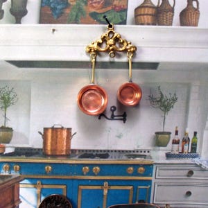 May include: Miniature copper cookware set hanging from a decorative gold-colored bracket. The set includes two small pans. The background features a blue and white kitchen scene with a stove and other kitchen accessories.