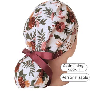 May include: A white floral scrub cap with a burgundy ribbon. The cap features a pattern of burgundy, peach, and pink roses with green leaves. The text "Satin lining option" and "Personalizable" are also visible.