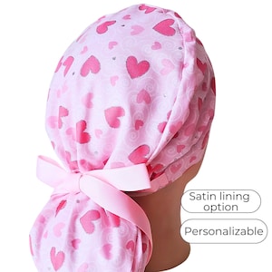 May include: A pink scrub cap featuring a pattern of pink hearts and a matching bow. The cap has a satin lining option and is personalizable. The cap is designed to be worn on the head.