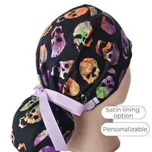 May include: Black skull print scrub cap with a light purple ribbon. The cap features a satin lining option and is personalizable. The skull design includes orange, purple, and green hues.