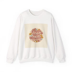 May include: White sweatshirt with a cream-coloured square graphic. The graphic features the words "Love You Tons" in a retro font, surrounded by hearts and dots in shades of red, orange, and blue. A cosy and stylish top.