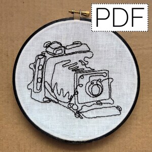 May include: Black and white line drawing of a vintage camera embroidered on a white fabric stretched over a round wooden hoop.