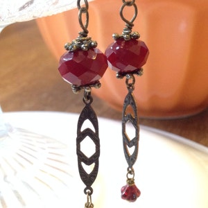 May include: A pair of dangle earrings with a dark red faceted glass bead and a decorative bronze pendant. The pendant has a geometric design.