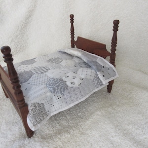 Pode incluir: Uma cama de boneca de madeira com uma colcha de retalhos branca e cinza. A cama tem uma cabeceira e um pé de cama com postes torneados.