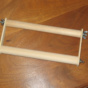 May include: A wooden weaving frame with two parallel dowels held together by rectangular side pieces. Wing nuts secure the dowels. The frame is made of light-coloured wood.