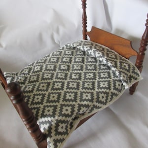 May include: A miniature wooden four-poster bed frame with a dark brown finish. The bed is dressed with a white and grey geometric patterned quilt and a matching pillow, featuring a repeating diamond-like design across the fabric surface.