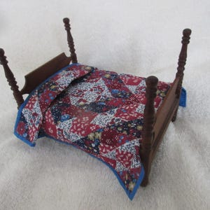 May include: Miniature wooden bed with a patchwork quilt. The bed frame is dark brown with four decorative posts. The quilt features a red, white, and blue floral pattern with a blue border. A charming piece for a dollhouse or miniature collection.
