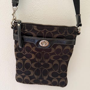 May include: A dark brown and black crossbody bag featuring a repeating logo pattern, a top zipper closure, and a front pocket with a silver-toned metal turn-lock clasp. The bag includes a black adjustable fabric strap attached with silver metal hardware.