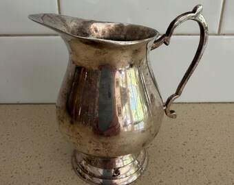 May include: A silver-toned metal pitcher featuring a curved handle, a wide pouring spout, and a rounded body that tapers toward a circular base. The surface of the pitcher shows signs of tarnishing and wear, with a reflective finish throughout.