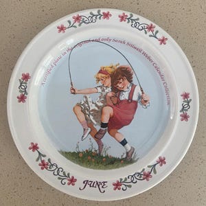 May include: A white ceramic plate with a blue center featuring two girls jumping rope. The plate has a pink floral border and the text "A certified plate in the original and only Sarah Stillwell Weber Calendar Collection." and "JUNE".