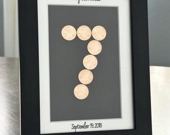 May include: A framed artwork featuring seven pennies arranged in the shape of the number seven. The black frame surrounds a white mat and a dark gray background. The names "Mickey and Rhonda" and the date "September 19, 2018" are written in script.