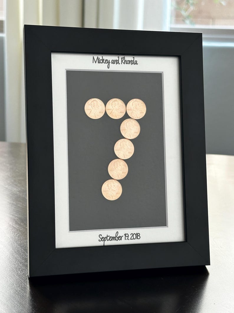 May include: A black rectangular picture frame containing a dark gray mat with a white border. The center features copper-colored pennies arranged in the shape of the number seven. The text "Mickey and Rhonda" and "September 19, 2018" is printed.