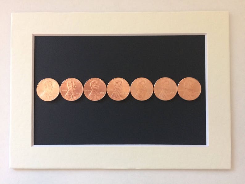 May include: A row of eight copper-coloured US pennies lined up on a black surface inside a white frame.