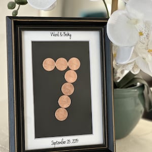May include: A black picture frame with gold trim containing a white mat board. The text Ward & Becky is printed at the top and September 28, 2019 at the bottom. A number seven is formed by six copper-colored coins on a black background.