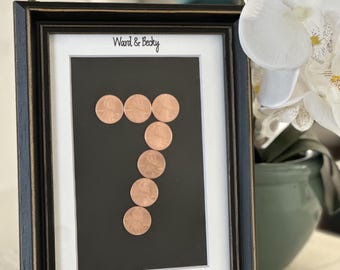 May include: A black picture frame with gold trim containing a white mount. The text Ward & Becky is printed at the top and September 28, 2019 at the bottom. A number seven is formed by six copper-coloured coins on a black background.