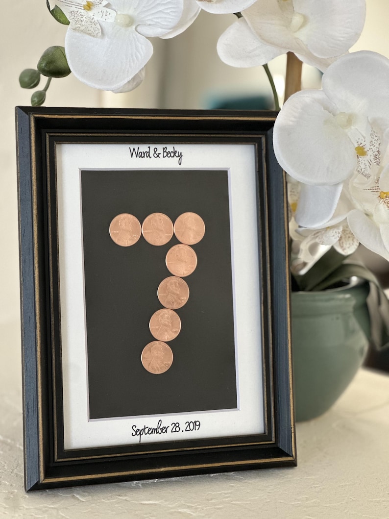 May include: A black picture frame with gold trim containing a white mat board. The text Ward & Becky is printed at the top and September 28, 2019 at the bottom. A number seven is formed by six copper-colored coins on a black background.