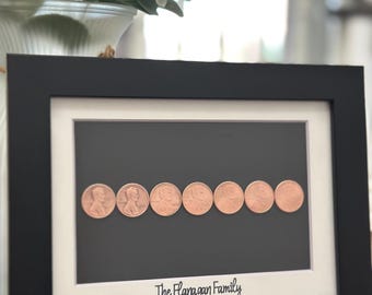 May include: A black rectangular frame with a white mat containing seven copper-colored pennies arranged in a horizontal row against a dark gray background. The white mat features the black printed text The Flanagan Family at the bottom center.