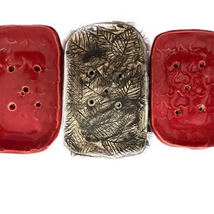 May include: Three rectangular ceramic soap dishes arranged side by side. The two outer dishes are a glossy, vibrant red with a subtle embossed heart pattern and five drainage holes. The center dish is metallic silver with a detailed leaf imprint.