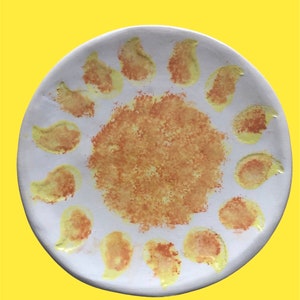 Puede incluir: Un plato de cerámica blanco con un diseño de sol amarillo y naranja. El sol está formado por un círculo con un centro texturizado y 12 formas de lágrima amarillas y naranjas alrededor del exterior.