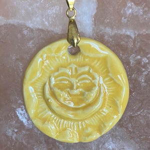 Pode incluir: Um pingente circular de cerâmica amarela com um relevo de um rosto de sol com raios radiantes. O pingente tem um acabamento brilhante com detalhes de rachaduras finas e está preso a um suporte de metal dourado em um colar de cordão preto.