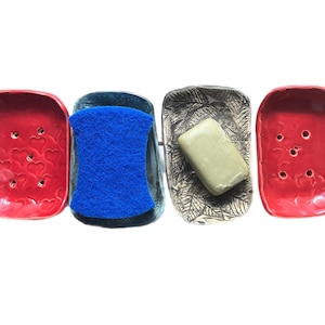 May include: Four rectangular soap dishes arranged in a row. Two are bright red with heart-shaped indentations and drainage holes. One is dark blue with a bright blue scouring sponge. One is grey with a leaf pattern and a rectangular bar of soap.