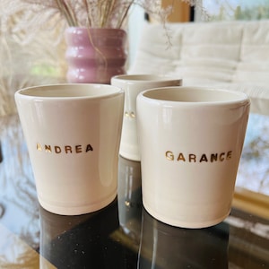 May include: Three off-white ceramic cups with gold lettering. The cups are labeled "ANDREA" and "GARANCE". The cups are on a reflective black and gold surface. A pink vase with dried flowers is in the background.