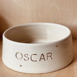 Op de afbeelding: Crèmekleurige keramische voerbak voor huisdieren met de naam "OSCAR" in gouden letters. De kom heeft een breed, ondiep ontwerp en is versierd met kleine gouden hartaccenten. De kom is ontworpen voor huisdieren.