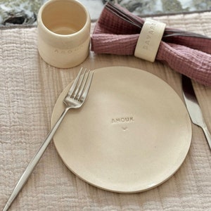Peut inclure: Une assiette en céramique blanche avec le mot "Anouk" en relief. Une fourchette en argent est placée sur l'assiette. Un anneau de serviette avec le mot "Bavard" est sur une serviette rose.