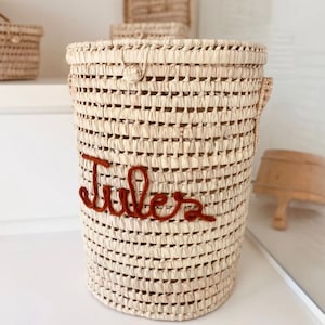 May include: Woven basket with the name "Jules" embroidered in a reddish-brown thread. The basket is cylindrical with a lid and handles. The basket is made of a light beige material.