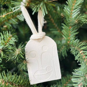 Peut inclure: Une décoration de Noël en feutre blanc avec un motif en relief de la scène de la nativité. L'ornement est suspendu à un ruban blanc.