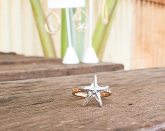 May include: A silver starfish ring with a gold band. The ring is sitting on a wooden surface.