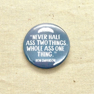 May include: A round, blue button with white text that reads "NEVER HALF ASS TWO THINGS. WHOLE ASS ONE THING." The text "RON SWANSON" is below the quote. The button is on a tan, textured surface.