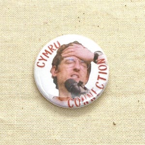 May include: A round, white button with a picture of a man with his hand on his forehead and a microphone. The button has the words "CYMRU CONNECTION" in red text around the image.