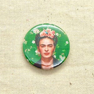 May include: A circular badge displaying a portrait of Frida Kahlo, adorned with a floral crown, against a green backdrop with white floral patterns. The image highlights Kahlo's distinctive features.
