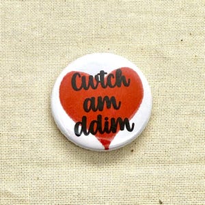 以下が含まれることがあります： 白いボタンに赤いハートのデザイン。ハートには黒い筆記体で「Cwtch am ddim」と書かれています。ボタンはベージュのテクスチャのある表面にあります。