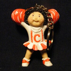 May include: A Cabbage Patch Kids cheerleader keychain. The doll is wearing a white and red cheerleader uniform with the letter "C" on the front. The doll has orange pom-poms and a gold keychain.