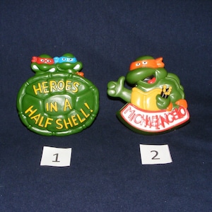 May include: Two Teenage Mutant Ninja Turtles toys. The first is a green turtle shell with the words "Heroes in a Half Shell!" The second is a green turtle with an orange mask and the word "Michelangelo" on the base.