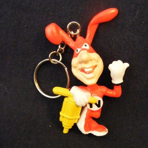May include: A novelty keychain featuring a cartoon rabbit character in red and white with a yellow tool. The rabbit has a smiling face and is holding the tool. The keychain has a gold-colored ring.