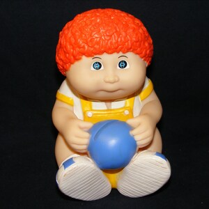 May include: A red-haired Cabbage Patch Kid doll wearing a yellow and white overall outfit and holding a blue ball.