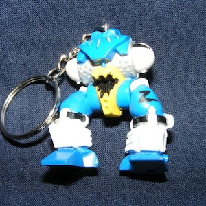 May include: Blue and white robot keychain with a yellow and black design. The robot is in a crouching position with its arms raised.