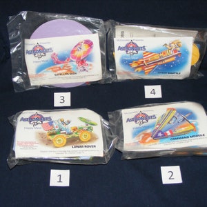 May include: Four sealed plastic bags containing McDonald's Astronauts Happy Meal toys. The toys are a satellite dish, a space shuttle, a lunar rover, and a command module. Each bag has a number on it, 1 through 4.