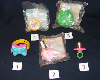 May include: Five plastic toys packaged in clear plastic bags. The toys are a yellow bracelet with a pink bow, a pink heart-shaped charm, a green ring with a pink flower, a yellow and pink toy with a spinning wheel, and a green toy with a red button.
