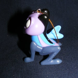 May include: A whimsical, stylized ornament in the shape of a creature with a purple head, black hair, and blue wings. The ornament has a gold string for hanging and is made of plastic. The creature's body is a mix of blue, purple, and gray.