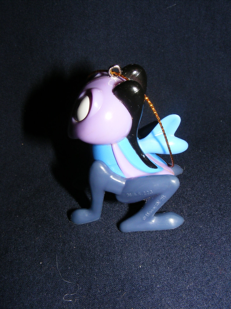 May include: A whimsical, stylized ornament in the shape of a creature with a purple head, black hair, and blue wings. The ornament has a gold string for hanging and is made of plastic. The creature's body is a mix of blue, purple, and gray.