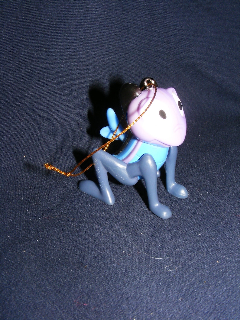 May include: A whimsical, kneeling figurine of a creature with a purple head, blue accents, and gray body. It has black eyes and a gold string for hanging. The figurine is a unique decorative item.